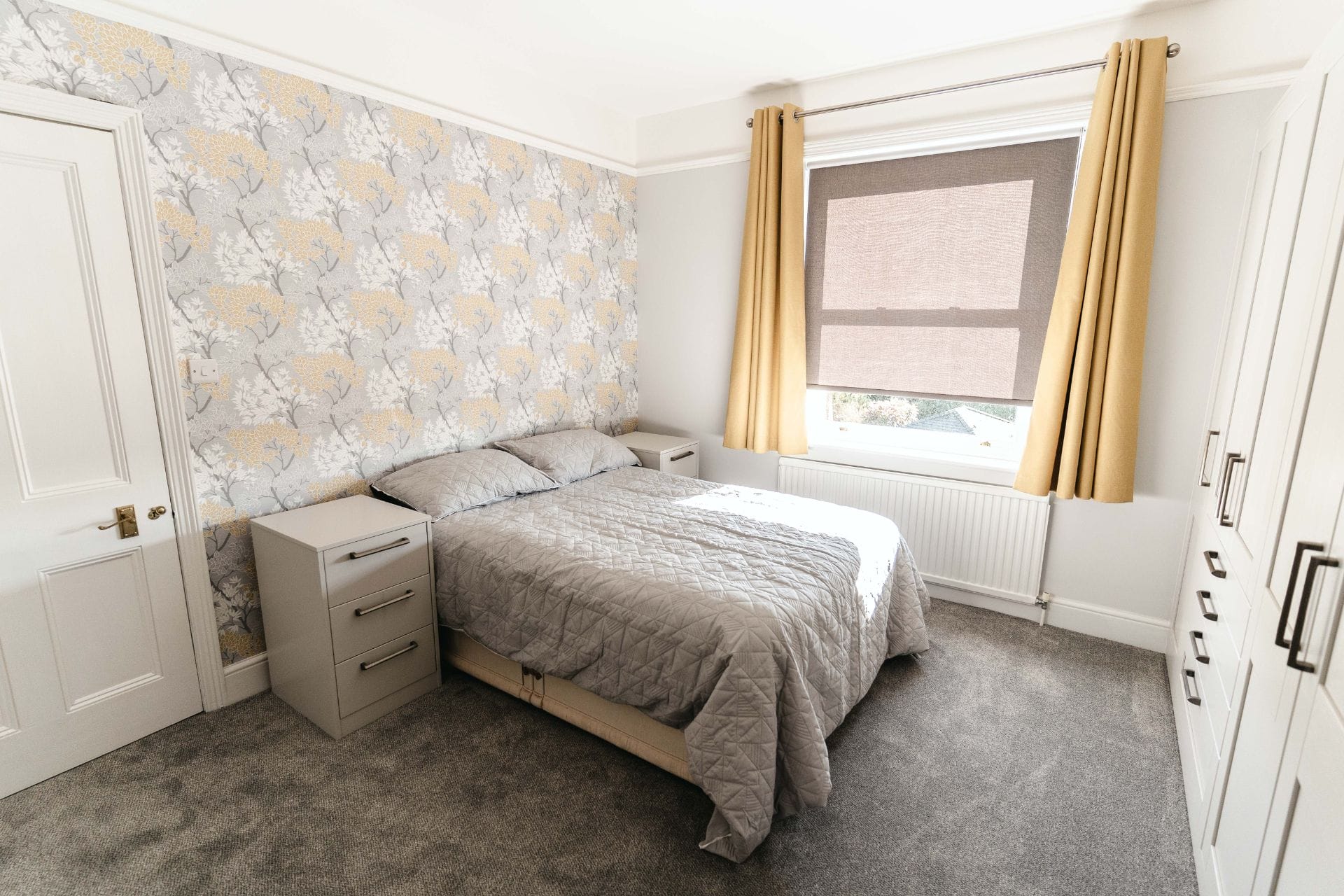 Traditional bedroom with floral feature wallpaper, grey fitted wardrobe, and yellow curtains