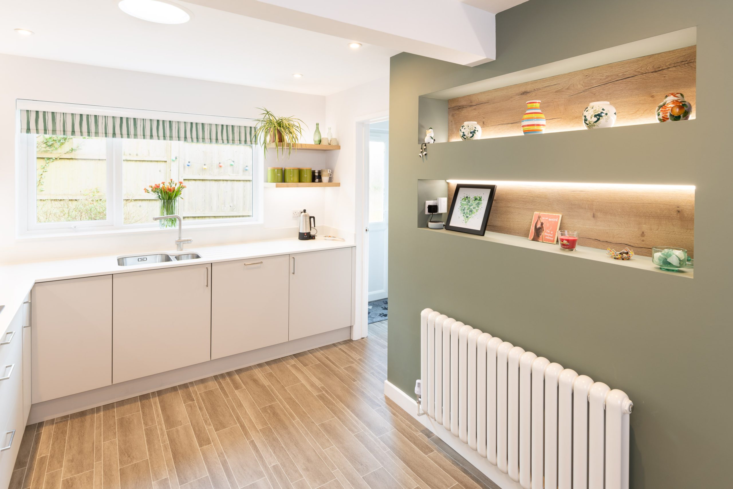 Open-plan kitchen with neutral cabinetry, white worktops, feature wall shelving and radiator by PB Kitchens & Bathrooms